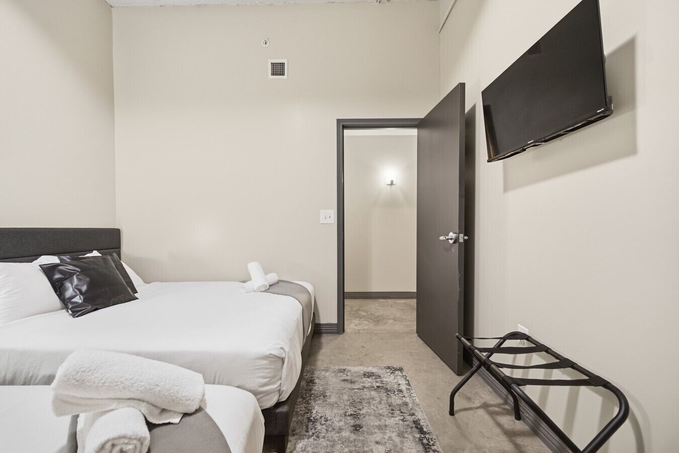 A hotel room with a bed, a TV mounted on the wall, a door leading to another room, and a small black metal rack.