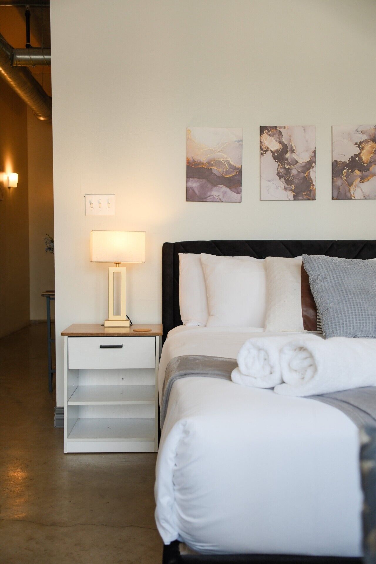 Habitación de hotel con cama, mesita de noche con lámpara y arte abstracto en la pared.