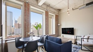 A modern living room with a large window, a sofa, a TV, and a dining area with chairs. - Stylish Downtown Stay | Pool, Gym, Valet Parking (Dallas)