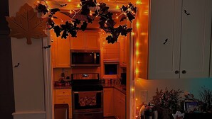 Eine Küche mit weißen Schränken, einer Mikrowelle und einem Herd. Dekorative Lampen und ein Halloween-Teppich sind vorhanden.