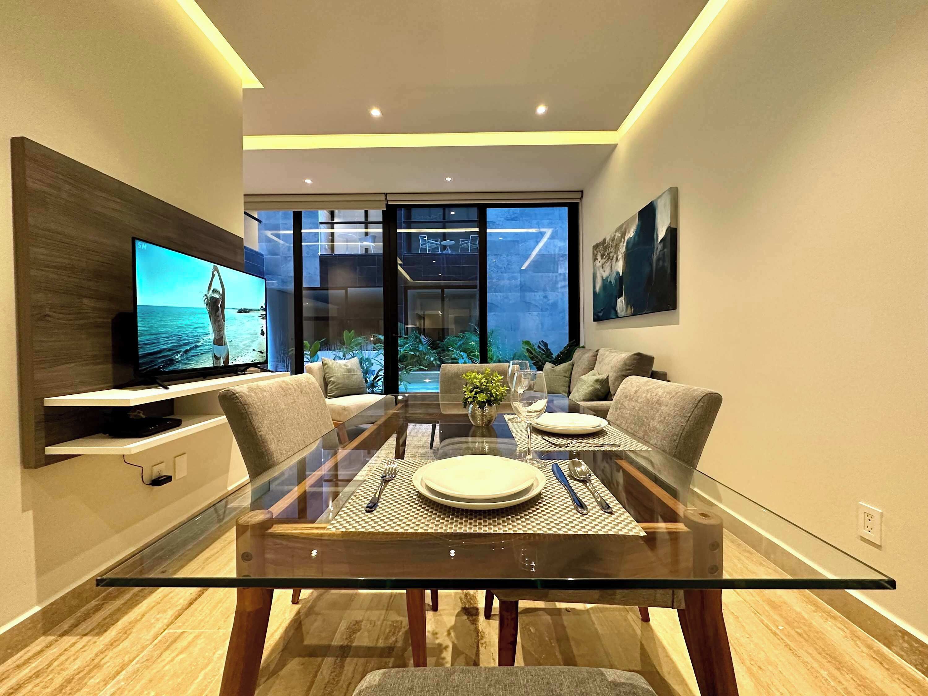 A modern living room with a glass dining table, sofas, a television, and a view of the outdoors.