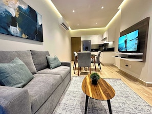A modern living room with a grey sofa, a wooden coffee table, and a flat-screen TV mounted on the wall. - Musa del Carmen · Garden Patio & Pool Access (Playa del Carmen)