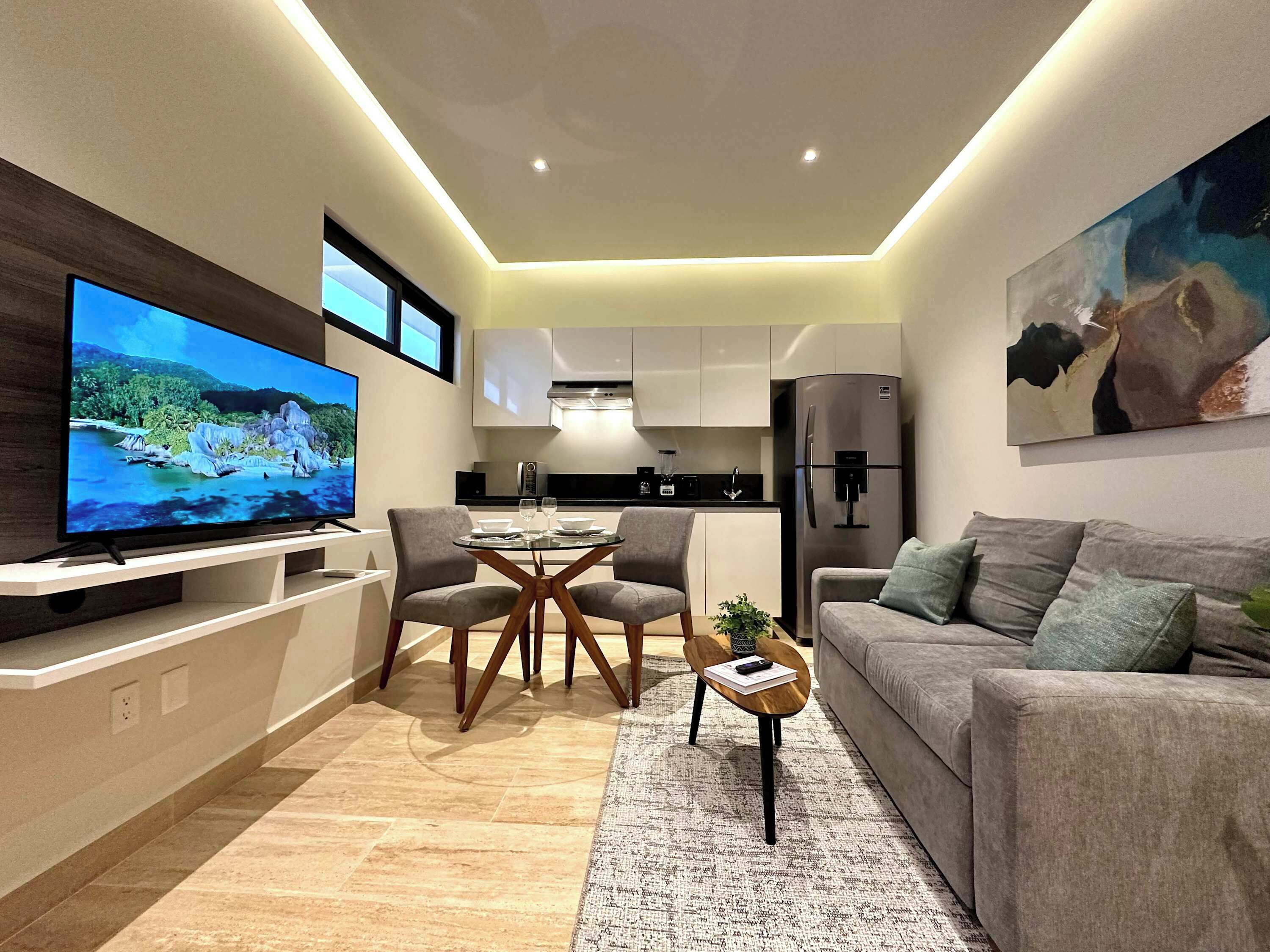 Ein modernes Wohnzimmer mit Flachbildfernseher, grauem Sofa und einem Essbereich mit Glastisch und Stühlen.