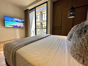 A hotel room with a bed, a television on the wall, a view of the outside balcony, and a hanging light fixture. - Musa del Carmen · 1BR w/Washer & Pool Access (Playa del Carmen)