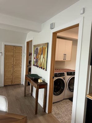 A laundry room with a washing machine and dryer, a wooden table with a mug, and a painting on the wall. - A little paradise for all seasons on the shores of Lake St-Alexis (Saint-Alexis-des-Monts)