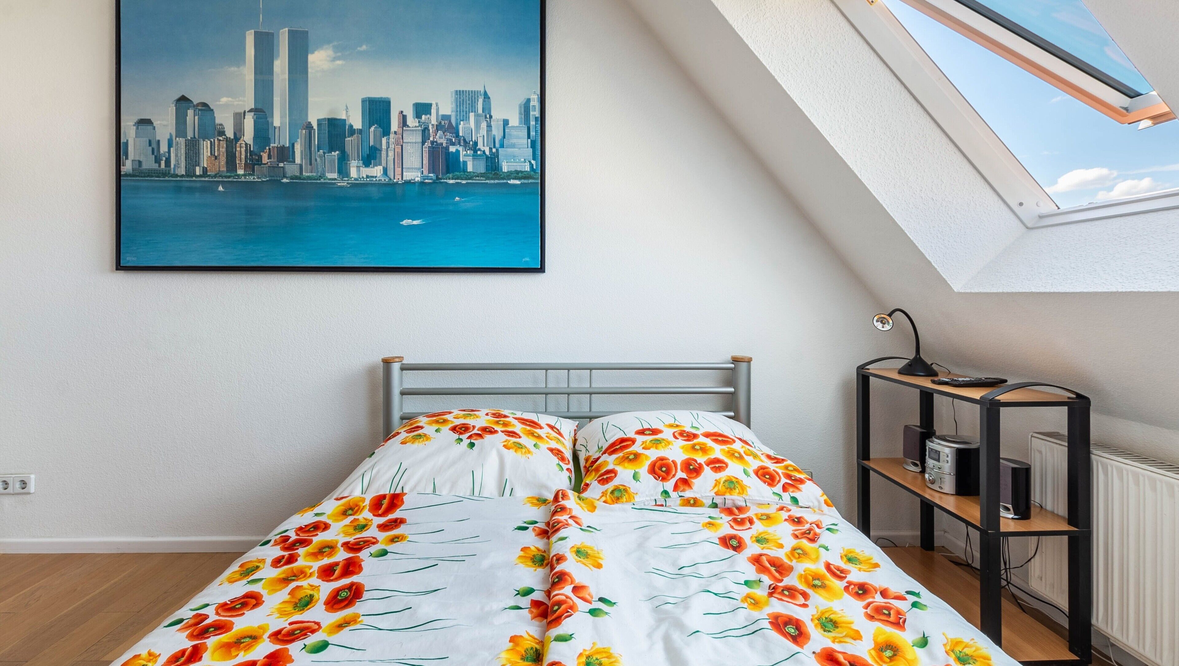 A bedroom with a bed, a cityscape painting, a skylight, and a desk with a lamp.