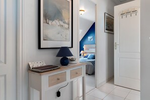 Um corredor com um quadro emoldurado, um abajur, uma mesinha com um livro e uma porta que dá para um quarto.