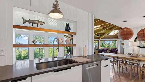 Eine moderne Küche mit integriertem Geschirrspüler, Spüle mit Armatur und Fenster mit Blick auf Bäume. Ein Essbereich mit Tisch und Stühlen, sowie eine Decke mit Holzbalken.