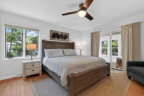 Une chambre à coucher avec un lit en bois, deux tables de chevet, un ventilateur de plafond et une vue sur l’extérieur grâce à de grandes fenêtres.