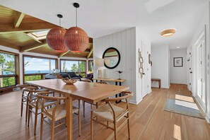 Un vaste espace dédié à la salle à manger, avec une table et des chaises en bois, de grandes fenêtres offrant une vue sur l’extérieur, et un plafond haut avec des poutres apparentes.