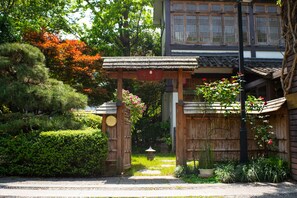 A traditional Japanese gate with a wooden structure, a stone lantern, and a pathway leading to a building with a tiled roof.