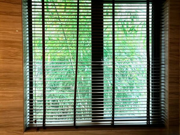 A window with horizontal blinds, a wooden frame, and a view of green foliage outside.