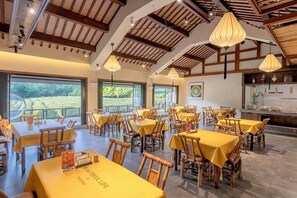 A restaurant with multiple tables and chairs, large windows, and a high ceiling with wooden beams.