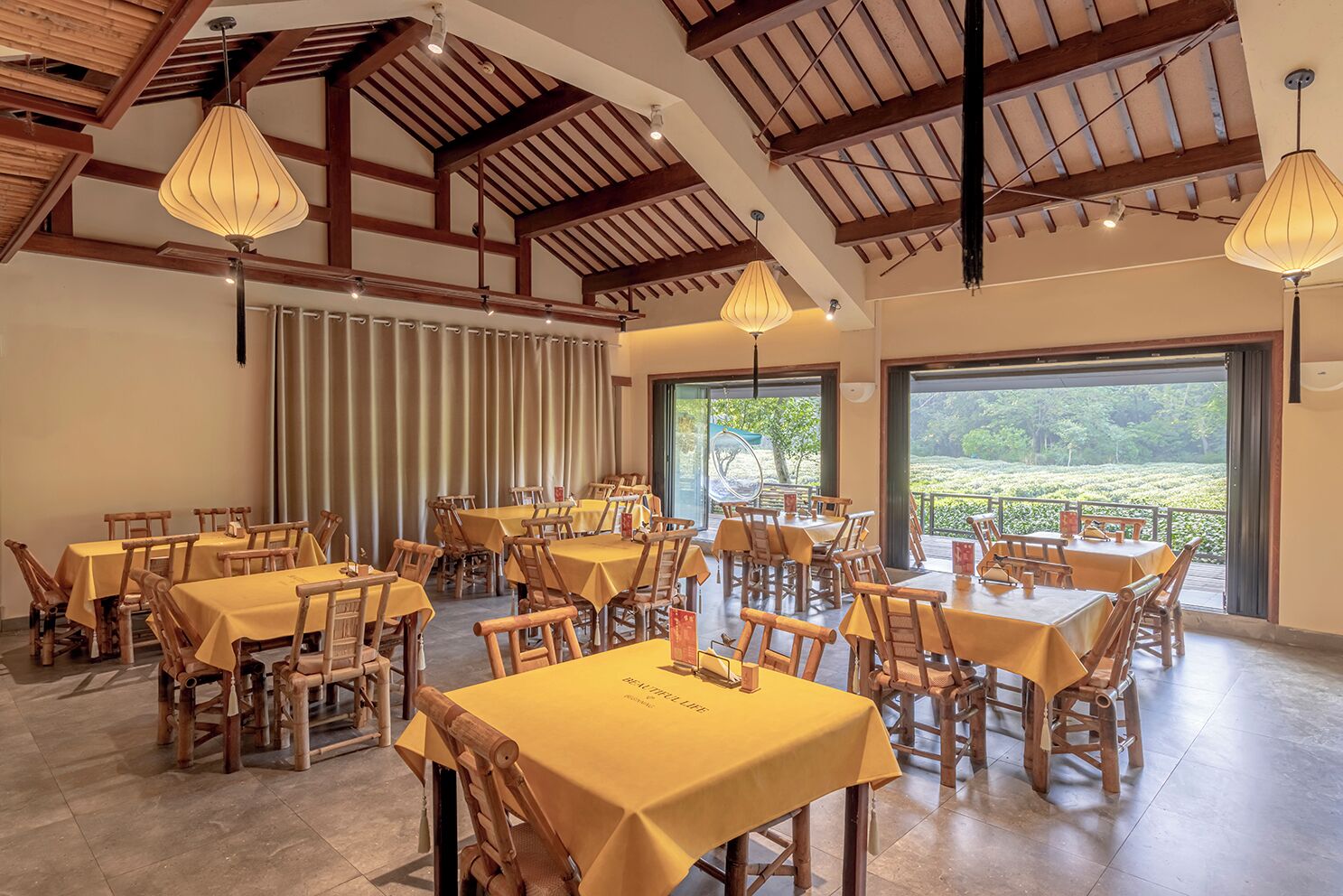 A dining area with multiple tables and chairs, wooden beams on the ceiling, and large windows offering a view of greenery.