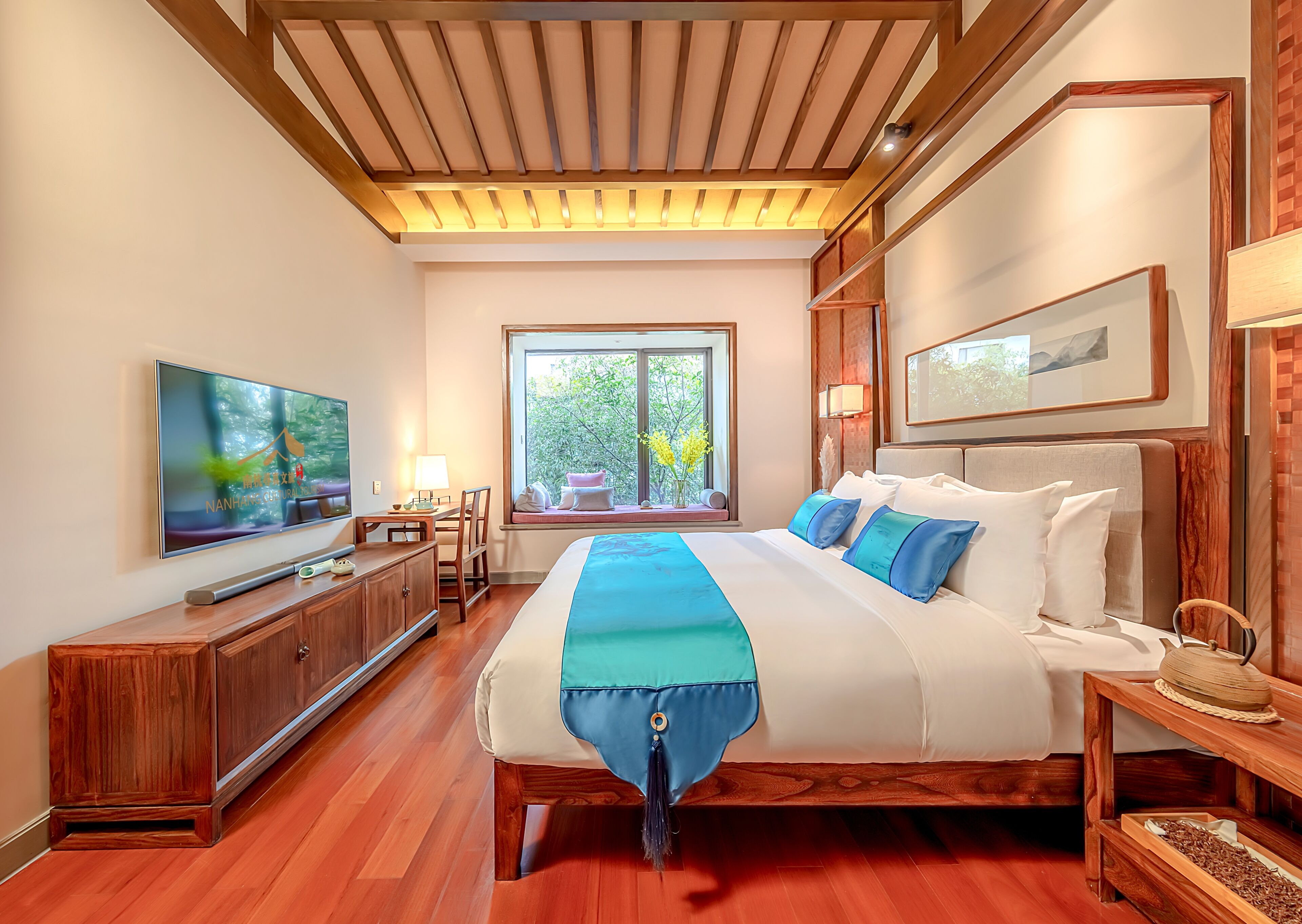A hotel room with a large bed, a TV, a desk, and a window with a view of trees.