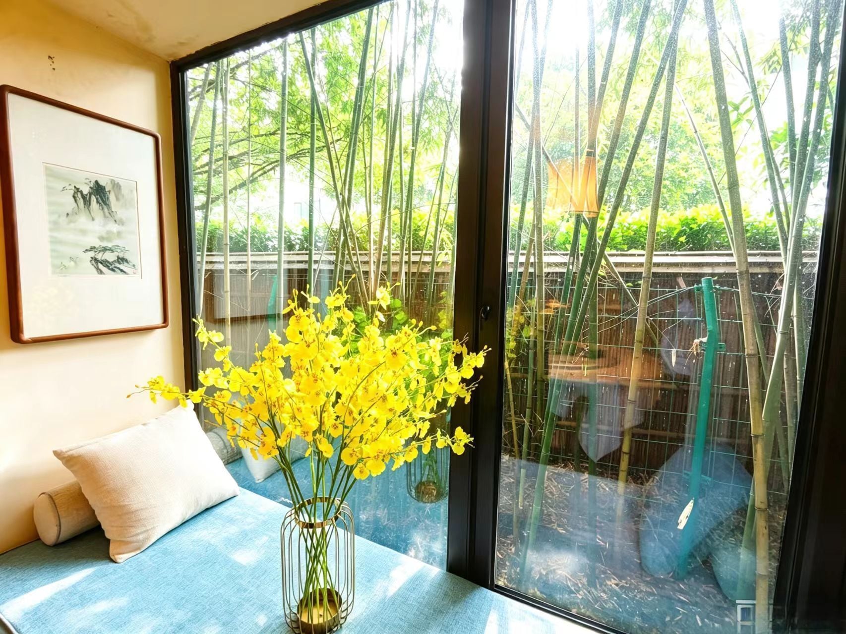 A room with a large window, a framed artwork on the wall, a vase with yellow flowers, and a blue sofa.