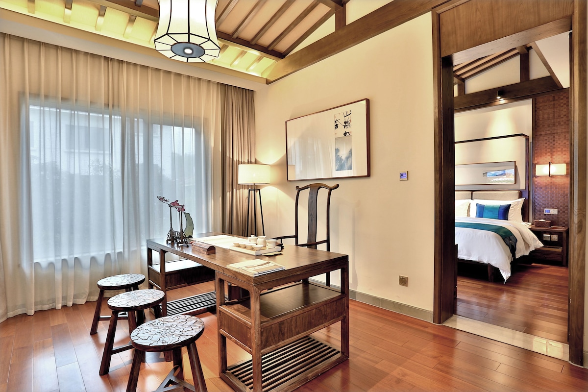 Une chambre d’hôtel avec une table à manger en bois, quatre chaises, une lampe, un lit avec un oreiller bleu et un tableau au mur.