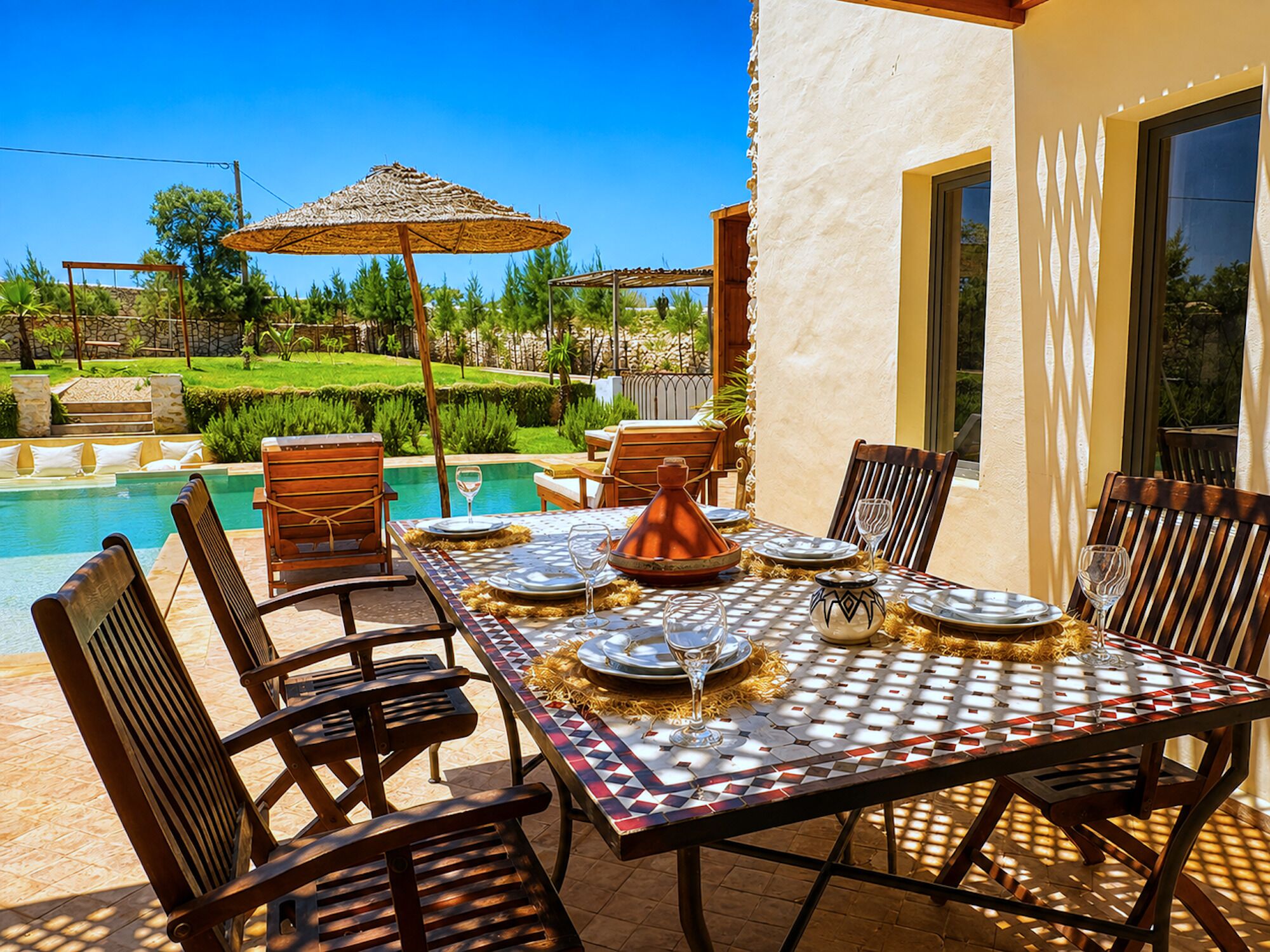 Un espacio exterior para comer con vista a la piscina, sillas de madera, una mesa con mantel estampado y un jarrón decorativo.