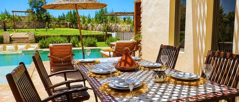 Un espacio exterior para comer con vista a la piscina, sillas de madera, una mesa con mantel estampado y un jarrón decorativo.