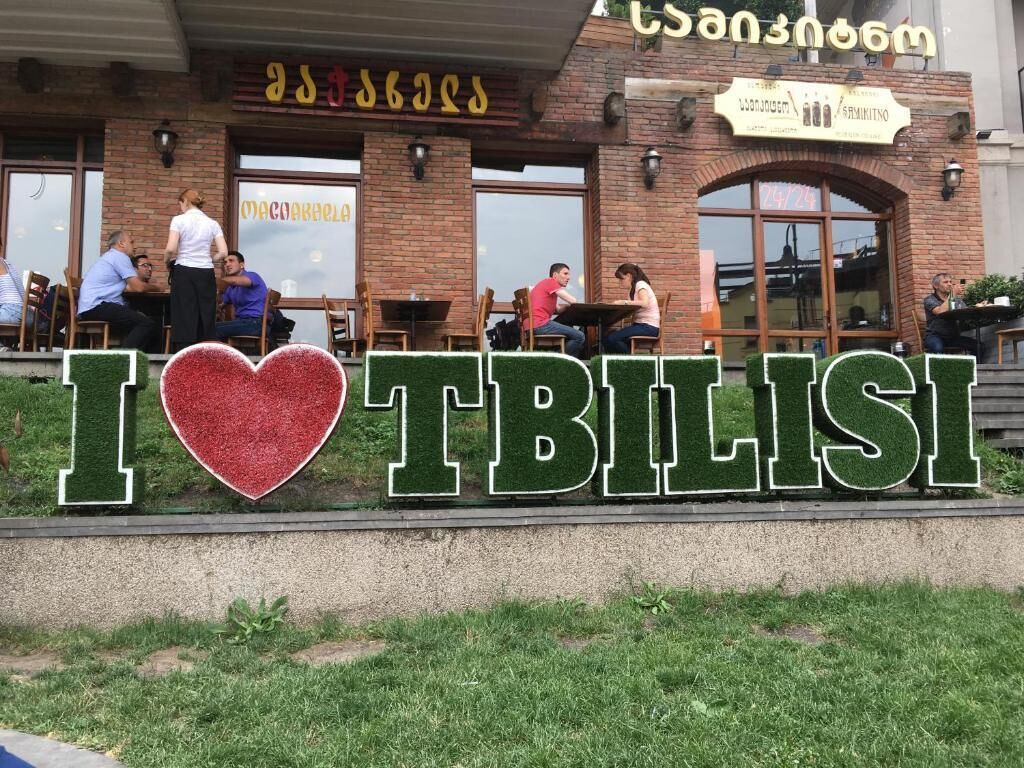 A sign with 'I ♥ Tbilisi' in front of a building with outdoor seating and a brick facade.