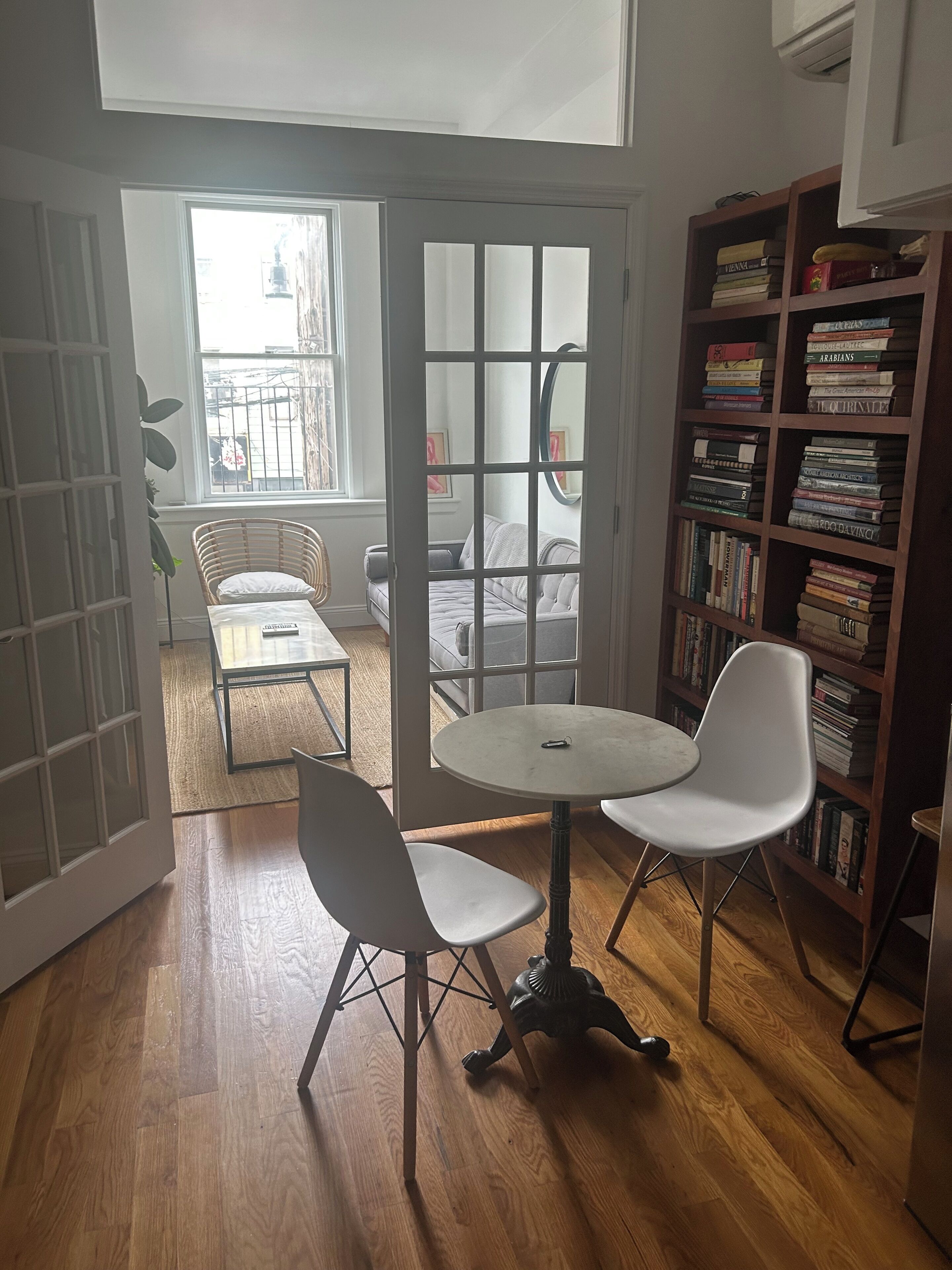 A room with a round table, two chairs, a bookshelf, and a view through a glass door.