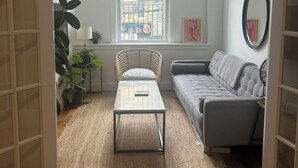 A living room with a sofa, a coffee table, a window with a view of buildings, and a potted plant.