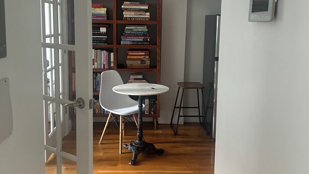 A room with a wooden floor, a white door, a white chair, a small round table, a bookshelf, and a wall-mounted device.