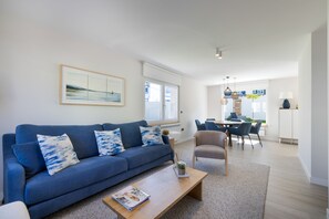 A modern living room with a blue sofa, a coffee table, and a dining area in the background. - Grandma’s House – Renovated Seaside Home in Central Muxía with Garden & Parking (Muxía)