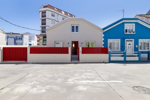 A building with a red gate, a white facade, and a blue house adjacent to it. - Grandma’s House – Renovated Seaside Home in Central Muxía with Garden & Parking (Muxía)