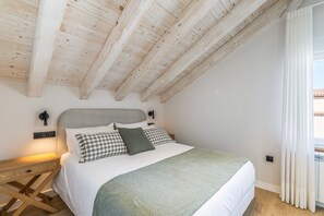 Ein Schlafzimmer mit schrägverlaufender Decke, einem Bett mit weißer Matratze und mehreren Kissen, einem Nachttisch mit Lampe und einem Fenster mit Vorhängen.