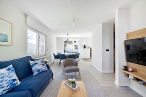 Ein modernes Wohnzimmer mit einer Couch, einem Couchtisch und einem Fernseher. Im Hintergrund befindet sich ein Essbereich mit Stühlen und einem Tisch.