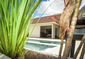 A pool area with a clear view of the pool, a covered outdoor seating area, and a building in the background.