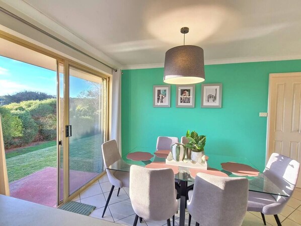 A dining area with a glass table and chairs, a hanging light fixture, and framed pictures on the wall.