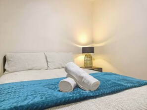 Een net opgemaakt bed met witte lakens en een blauwe deken, een wit kussen en een lamp met een draadframe lampenkap op een nachtkastje.