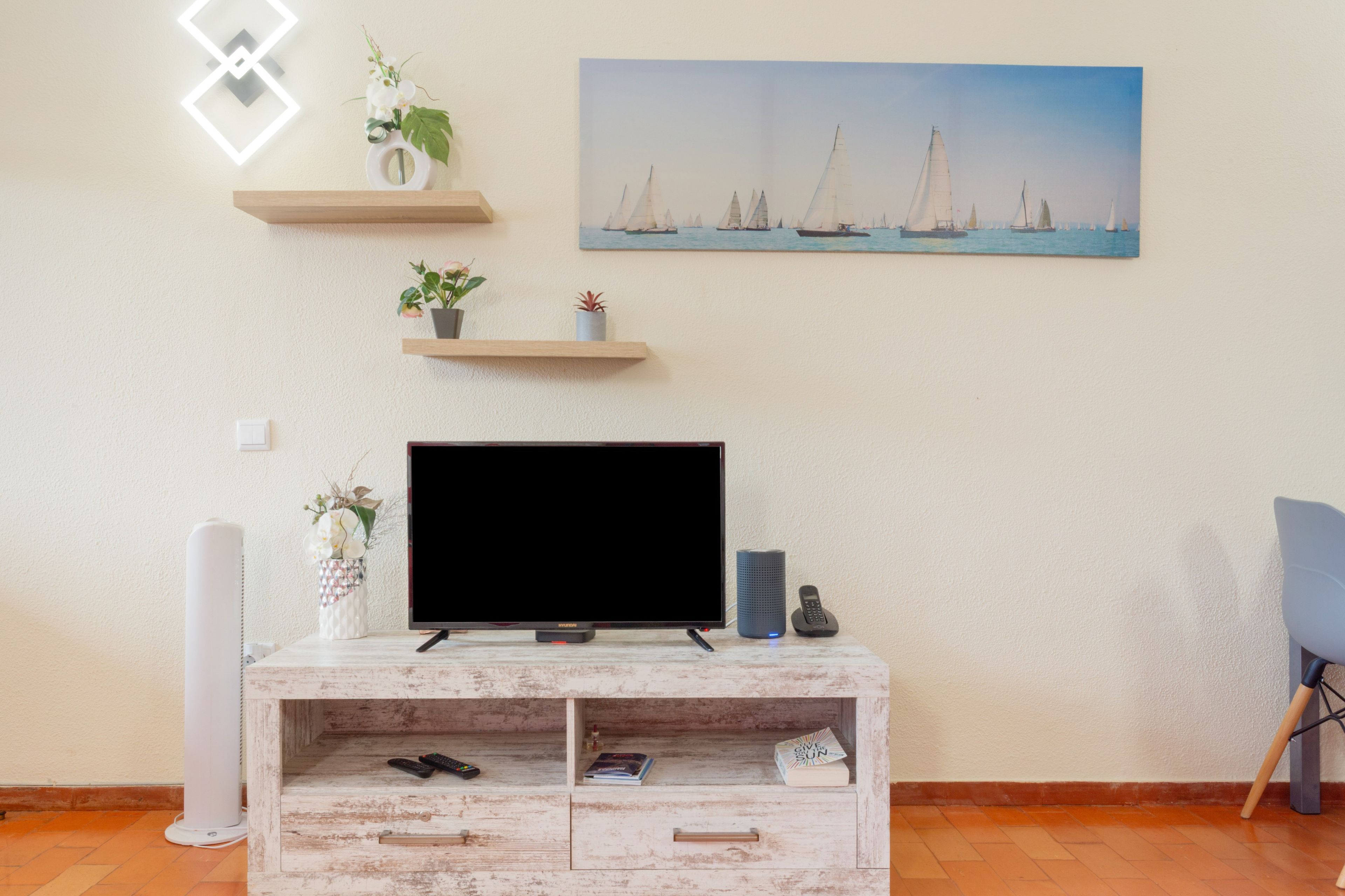 A living room with a flat-screen TV on a wooden TV stand, a painting of sailboats on the wall, and a small plant on a shelf.