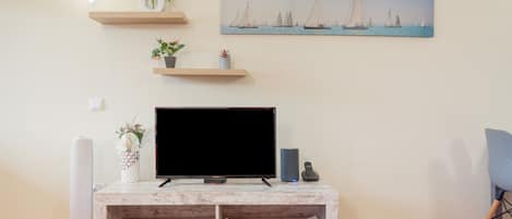 A living room with a flat-screen TV on a wooden TV stand, a painting of sailboats on the wall, and a small plant on a shelf.