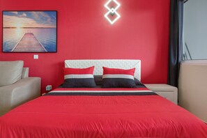 A hotel room with a red wall, a bed with a red and white striped bedspread, two pillows, a nightstand, a beige armchair, and a painting of a sunset over water. - Peaceful Retreat in Santa Cruz, Madeira — Steps from the Airport (Machico)