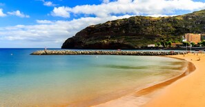 Ein Strand mit klarem Wasser, einem steinernen Wellenbrecher und Gebäuden im Hintergrund.