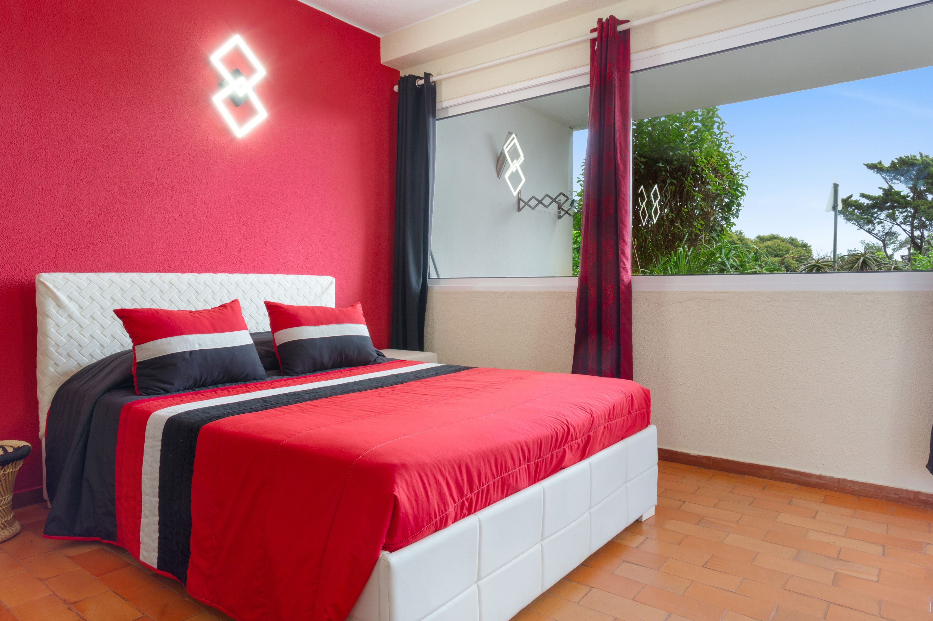 A bedroom with a bed, a window with curtains, and a balcony with a view of greenery.
