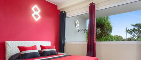 A bedroom with a bed, a window with curtains, and a balcony with a view of greenery.