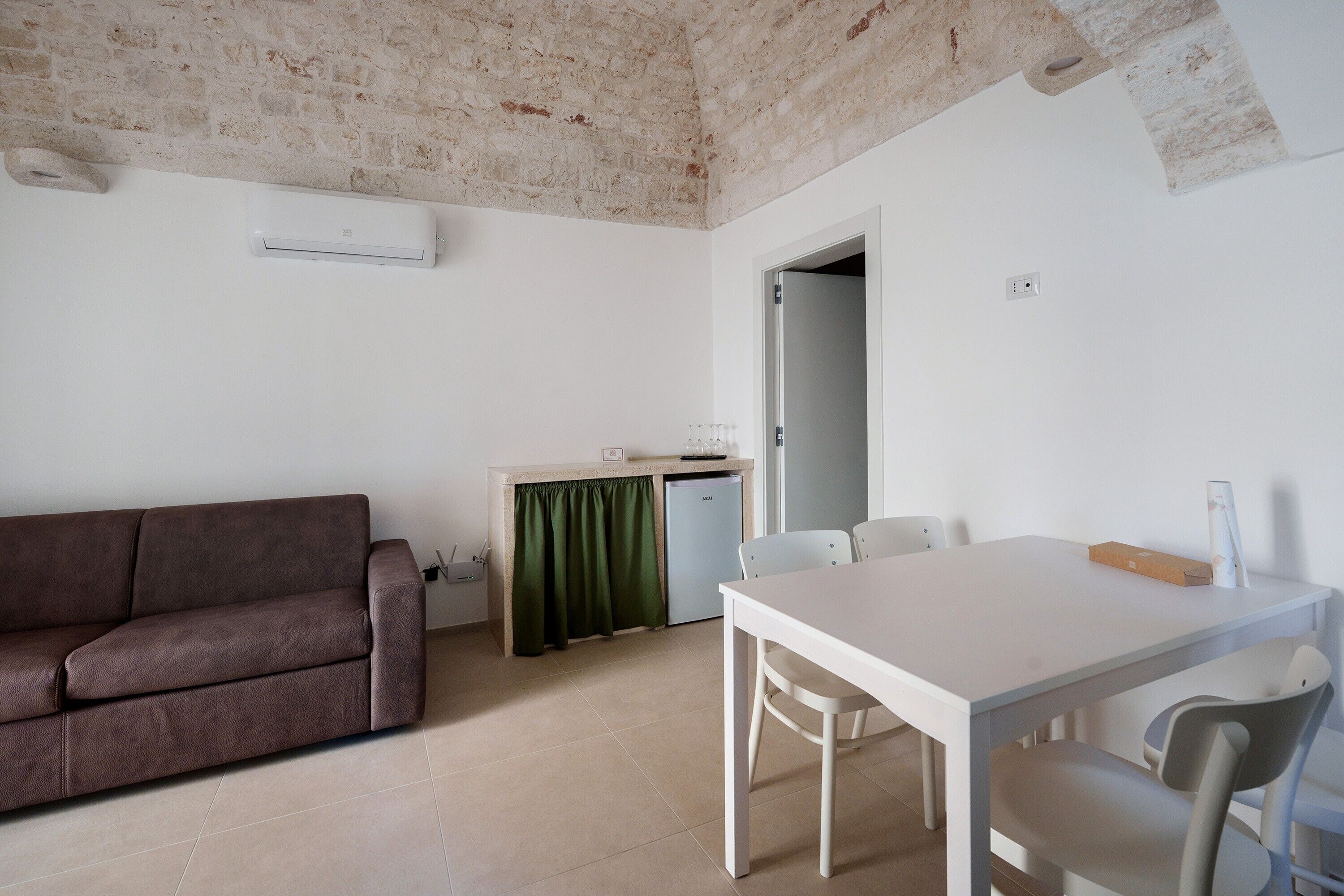 Una habitación con un sofá marrón, una mesa blanca con sillas, una kitchenette pequeña y una puerta que da a otra habitación.