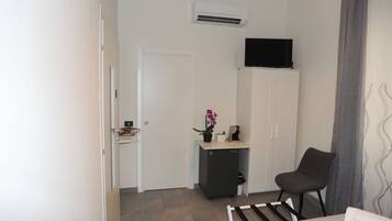 Une chambre d’hôtel avec un lit, une chaise, un bureau agrémenté d’une plante en pot, un téléviseur posé sur un meuble et un climatiseur fixé au mur.