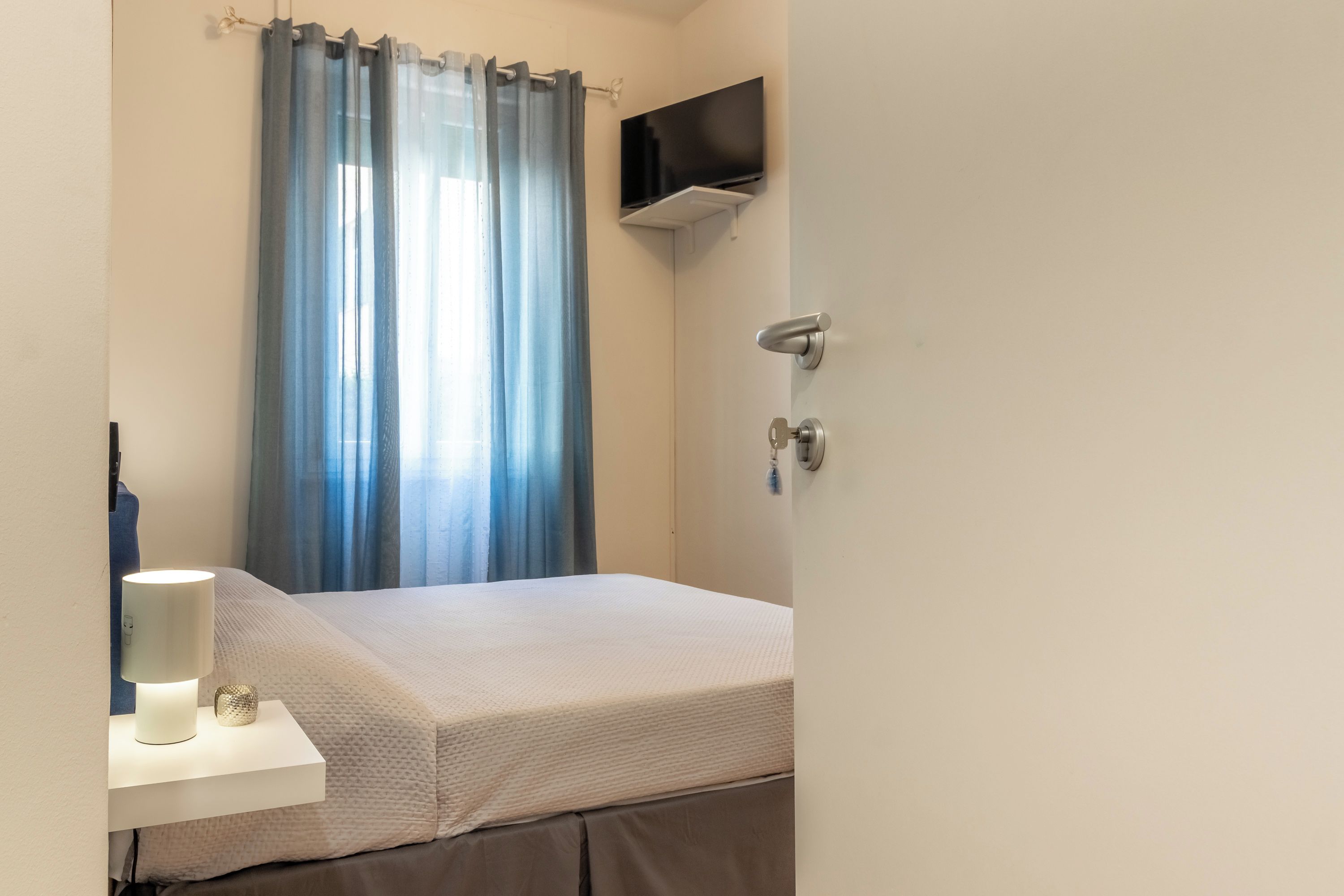 Una camera d'albergo con un letto, un comodino con una lampada, una TV a parete e una finestra con tende.
