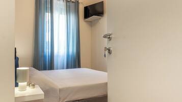 Una camera d'albergo con un letto, un comodino con una lampada, una TV a parete e una finestra con tende.