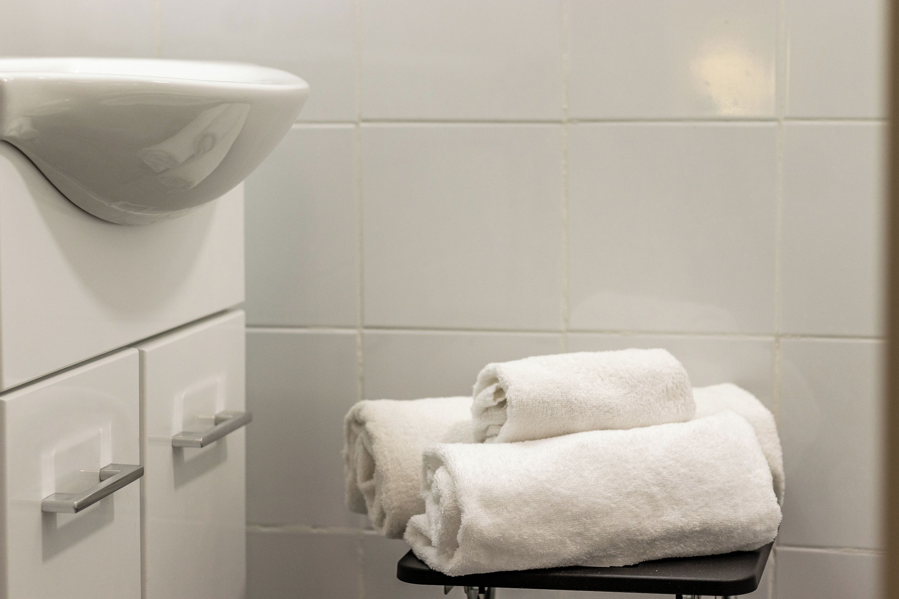 Une salle de bain avec un lavabo blanc, un porte-serviettes avec des serviettes pliées et des murs recouverts de carreaux blancs.
