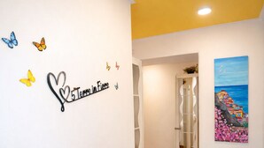 Un couloir orné d’une enseigne décorative, avec une plante en pot, une porte en verre donnant sur une salle de bain et un tableau accroché au mur.
