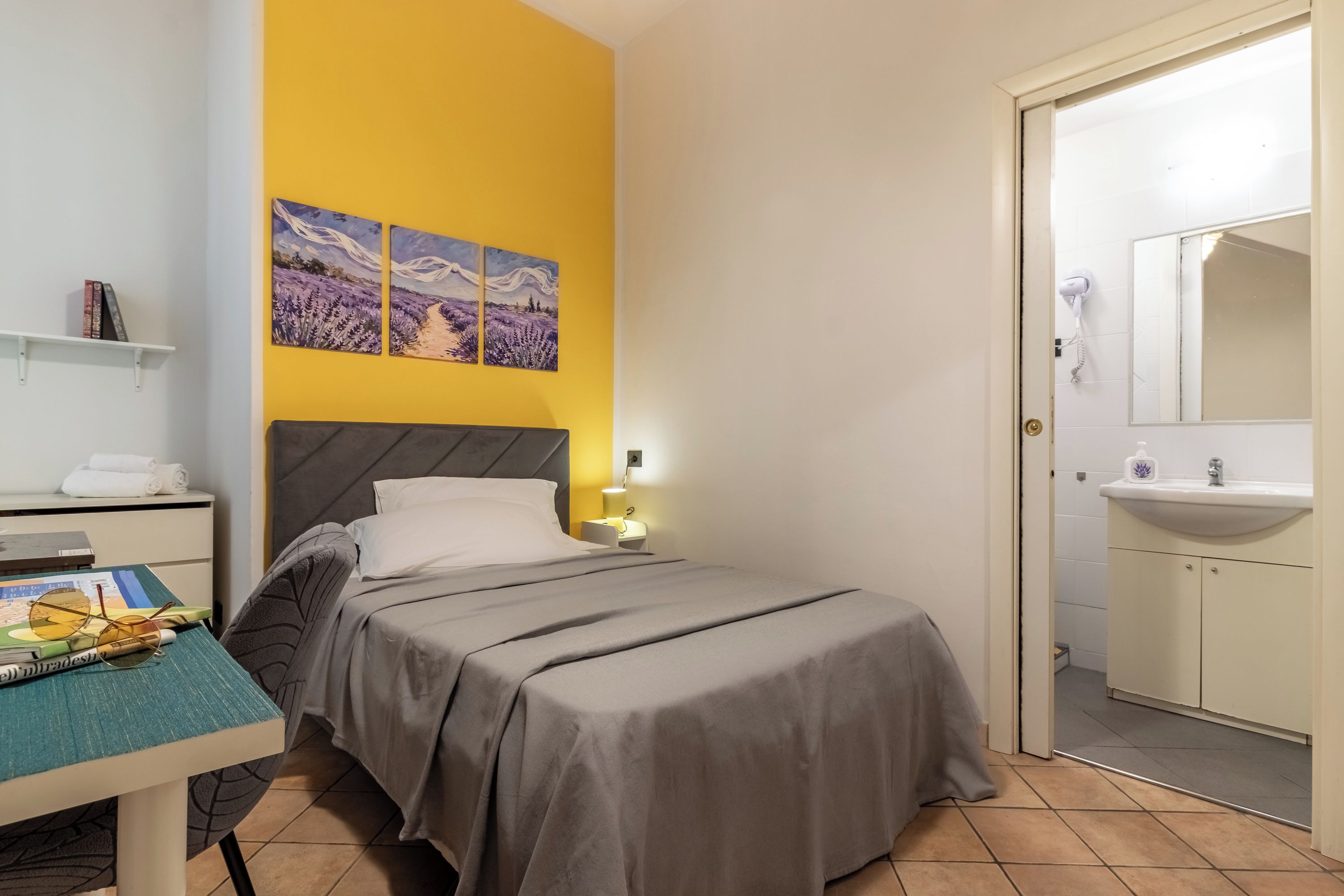 A bedroom with a bed, a nightstand, a lamp, a shelf with books, and a door leading to a bathroom.