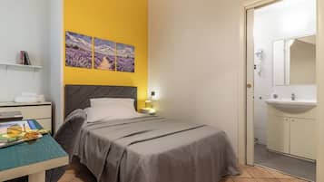 A bedroom with a bed, a nightstand, a lamp, a shelf with books, and a door leading to a bathroom.