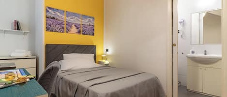 Un dormitorio con cama, mesita de noche, lámpara, estante con libros y puerta que da a un baño.