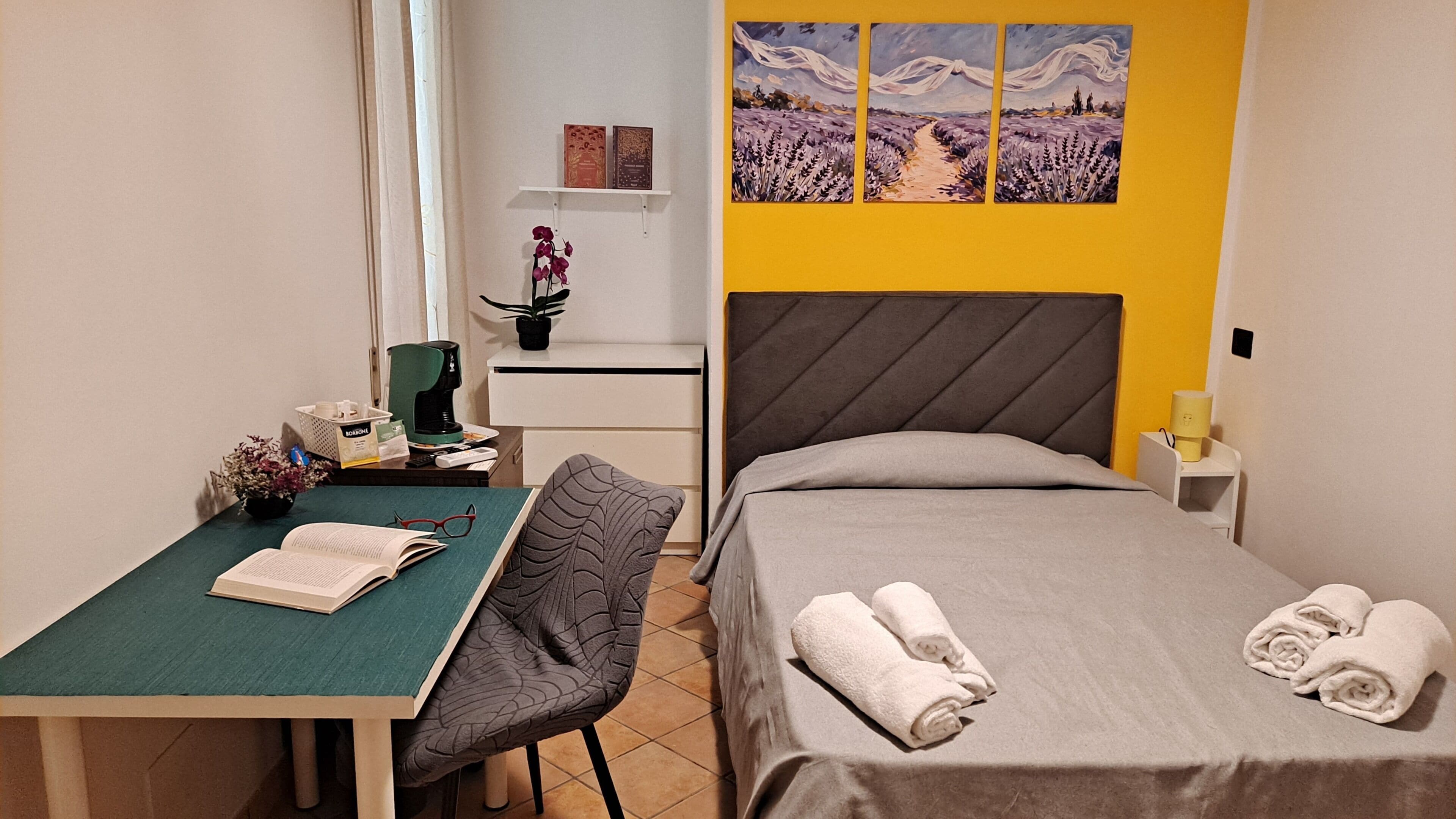 A hotel room with a bed, a desk, a chair, a coffee maker, a plant, and artwork on the wall.